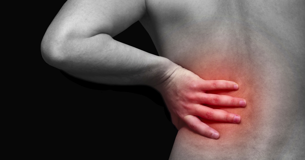 The image shows a close-up of a person’s back, highlighting a hand placed on the lower back, indicating pain or discomfort, with a dark background.