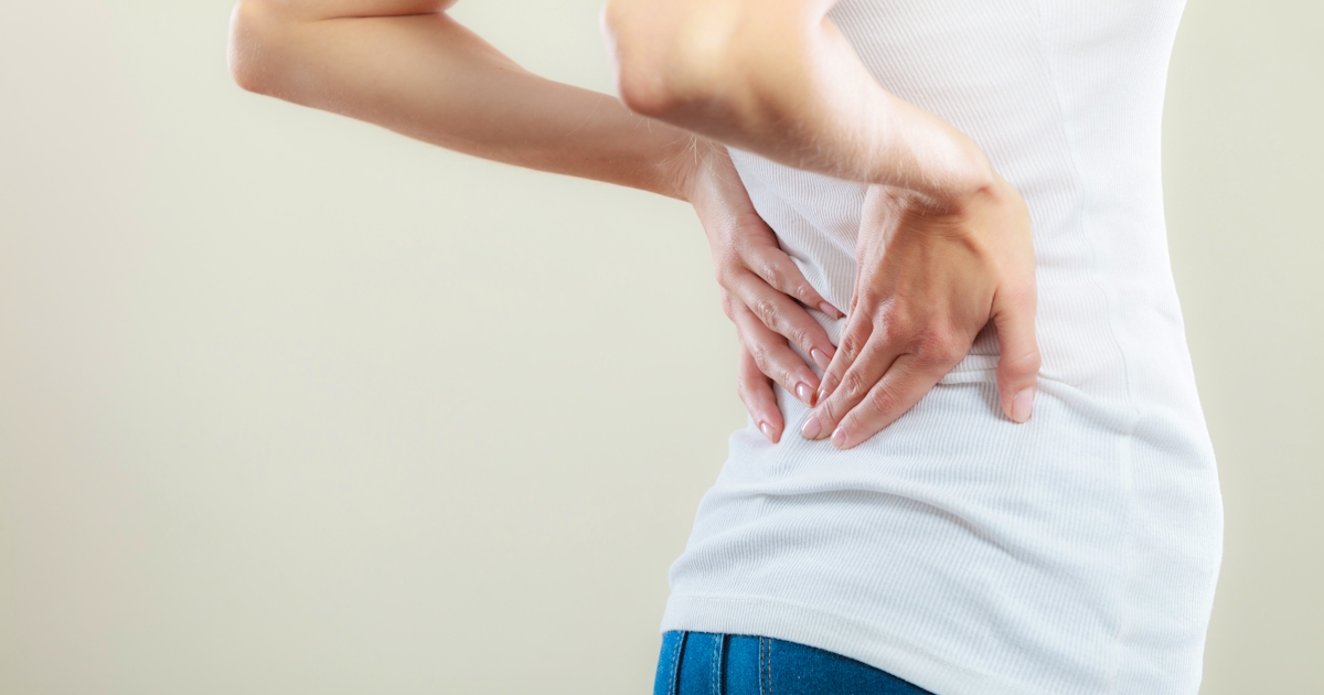 The image shows a person in a white shirt and jeans, experiencing discomfort in their lower back while gripping that area.