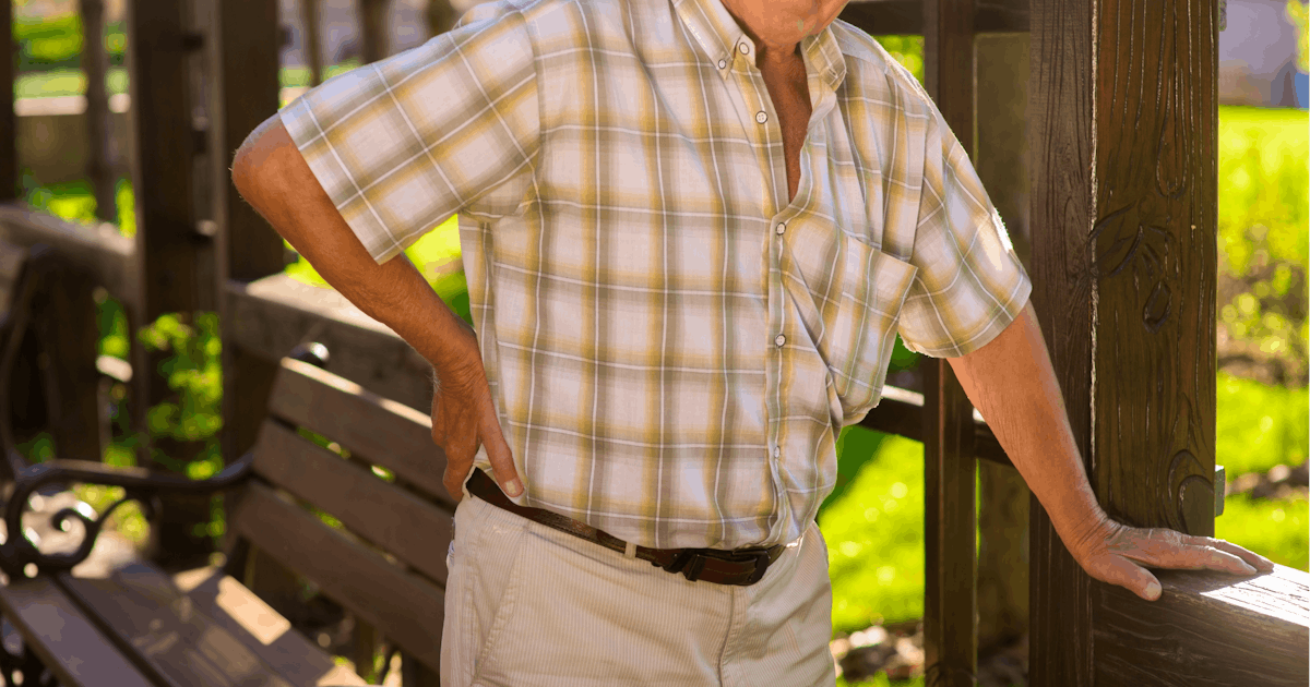 The image shows a middle-aged man in a plaid shirt and light pants, leaning against a railing in a park-like setting with green grass and benches.