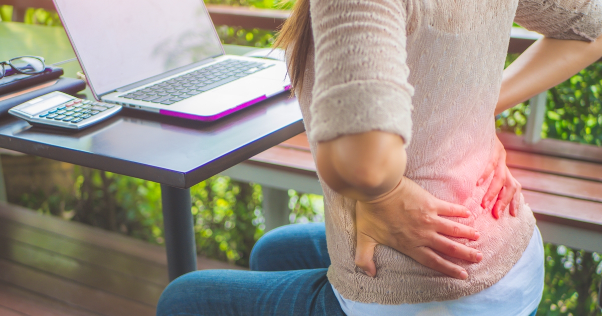 A person sitting at a table with a laptop appears to be experiencing back pain, holding their lower back with discomfort.