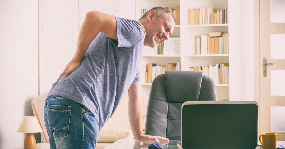 A man in a casual shirt is leaning over a desk, holding his lower back with a pained expression, in a bright office setting.