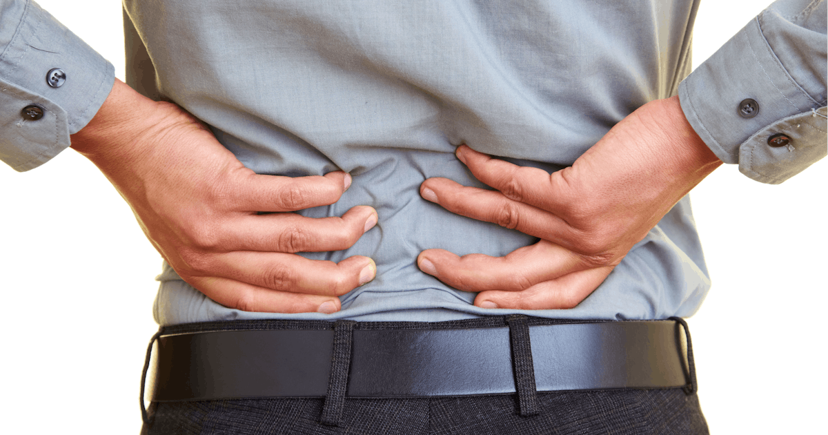 A person stands with their back facing the camera, holding their lower back with both hands, indicating discomfort or pain. They're wearing a gray shirt.