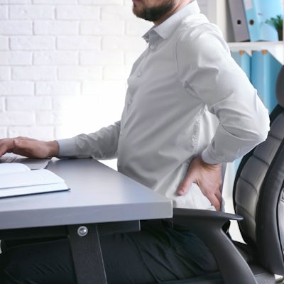 A person in a white shirt is sitting at a desk, looking at a laptop while holding their lower back, suggesting discomfort or pain.