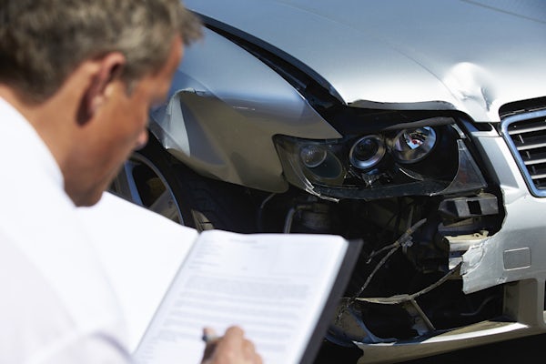 The image shows a person examining a damaged car, taking notes about the accident. The car has a noticeable dent on the front.