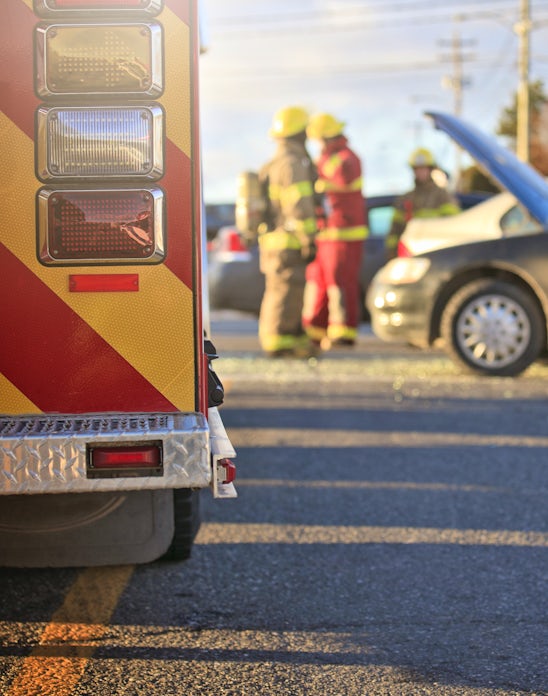 The image shows a fire truck with flashing lights, firefighters attending to a car accident, and a car with its hood raised on a city street.