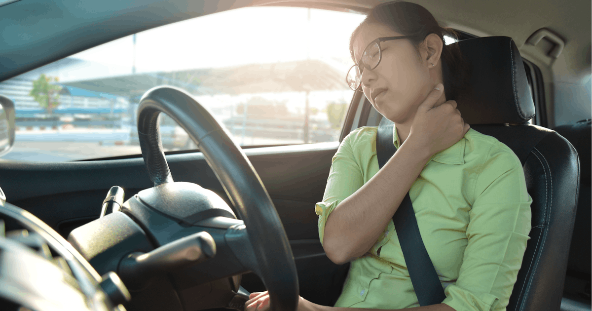 A woman sitting in a car grips the steering wheel, appearing to stretch her neck in discomfort, with sunlight streaming through the window.