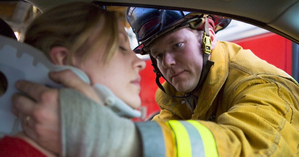 A firefighter is assisting a woman in a car, ensuring her safety with a neck brace after an incident. The scene focuses on their serious interaction.