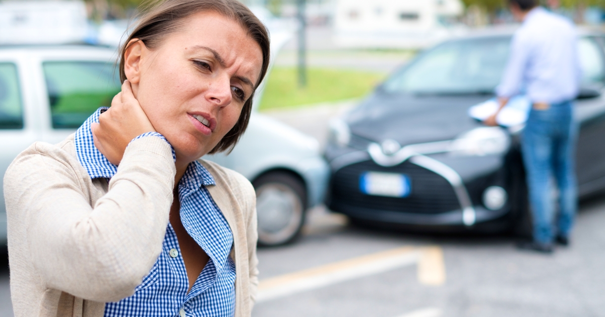 A woman is holding her neck, appearing to be in pain, while another person is inspecting a car involved in an accident nearby.