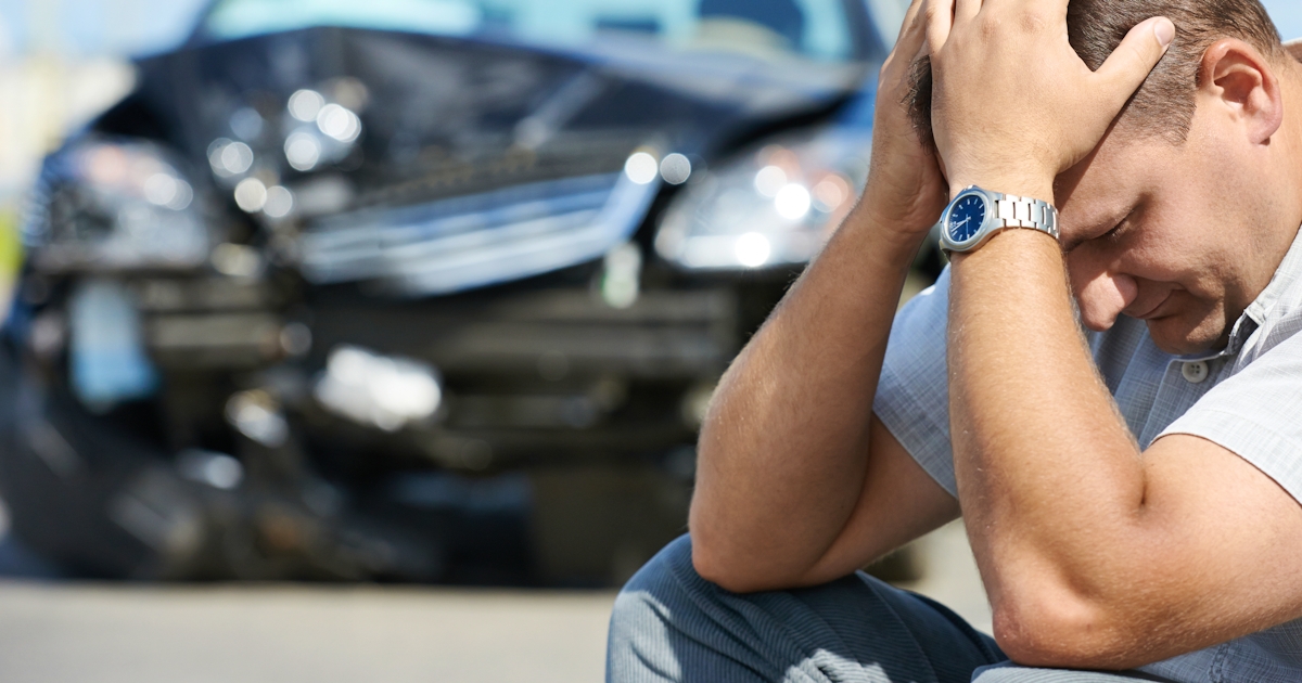 A man sits on the ground looking distressed, holding his head, with a damaged black car in the background, indicating a recent accident.