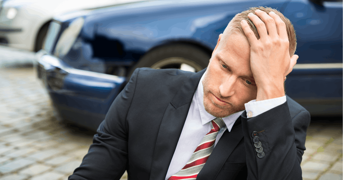Young man in suit with hand on head sits on ground in front of wrecked car