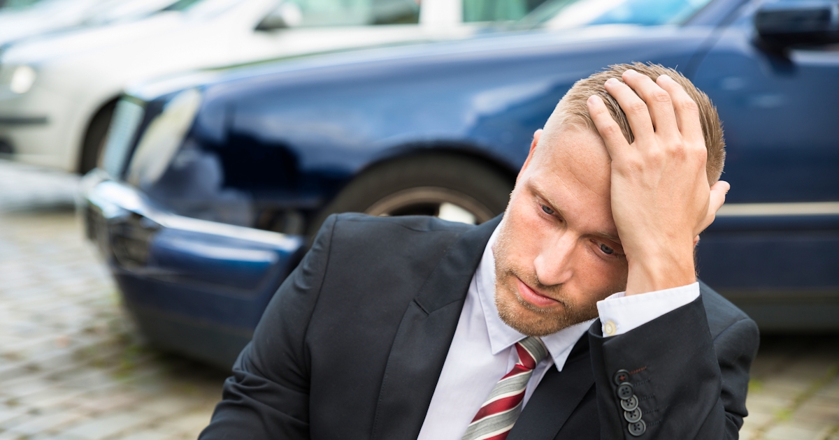 Young man in suit with hand on head sits on ground in front of wrecked car