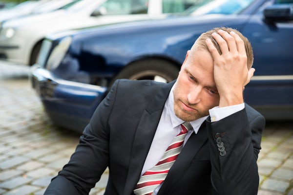 Young man in suit with hand on head sits on ground in front of wrecked car