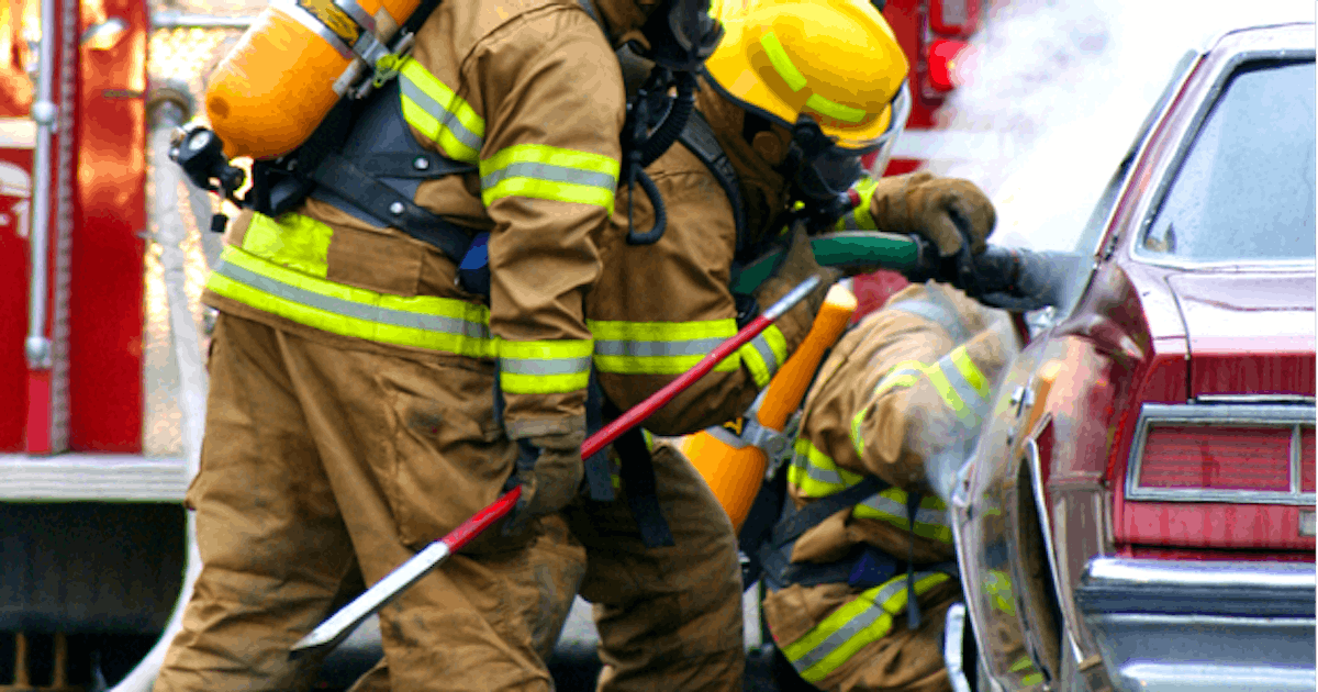 Firefighters in protective gear are extinguishing a fire in a car while a fire truck is visible in the background, with smoke rising from the vehicle.