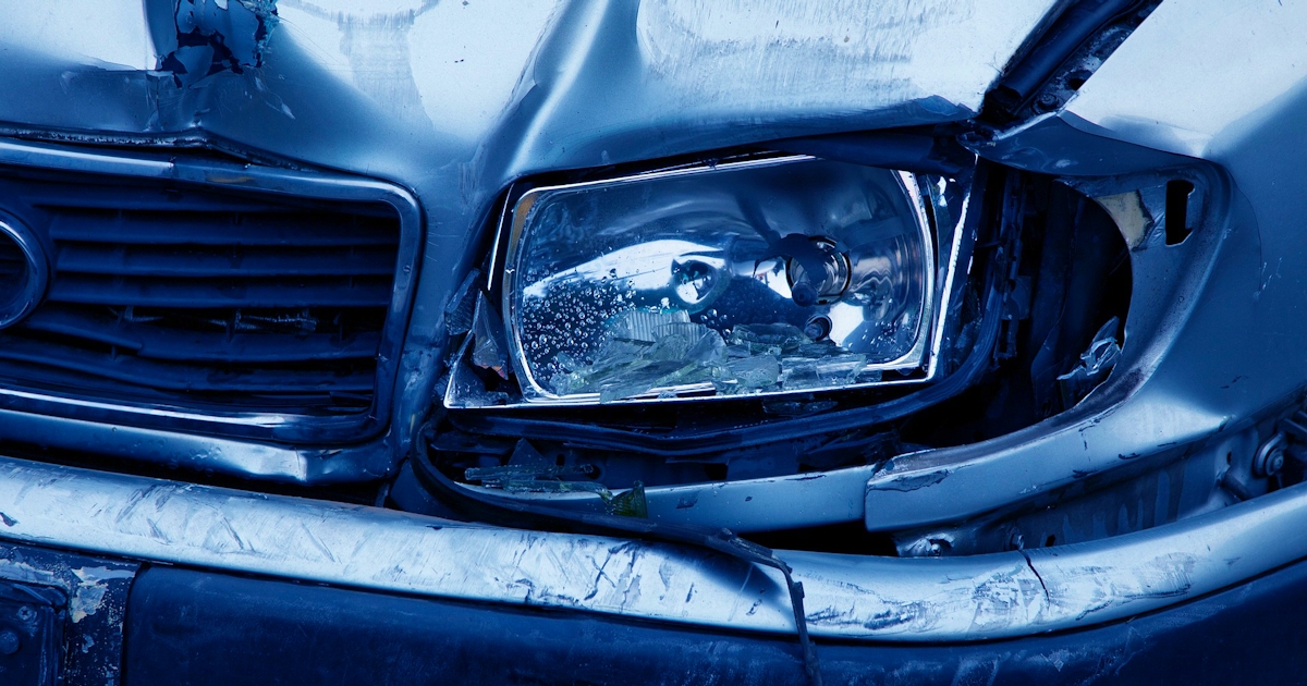 The image shows the front of a damaged car, with a crumpled hood and a broken headlight, highlighting the extent of the collision.