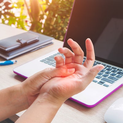 A person is holding their wrist in discomfort while seated at a desk with a laptop and some stationary items in the background.