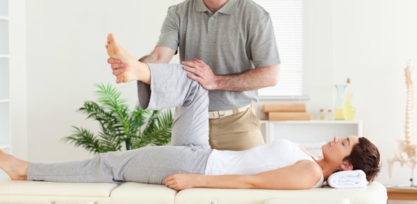 The image shows a man performing a leg stretch on a woman lying on a treatment table in a bright, serene therapy room.