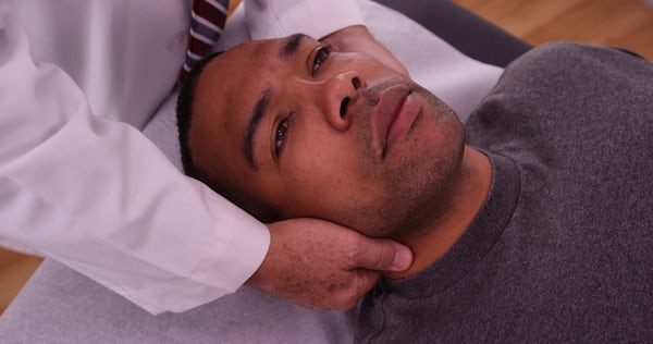 The image shows a man lying on a treatment table while a person in a white coat gently holds his head, possibly during a medical examination or therapy.