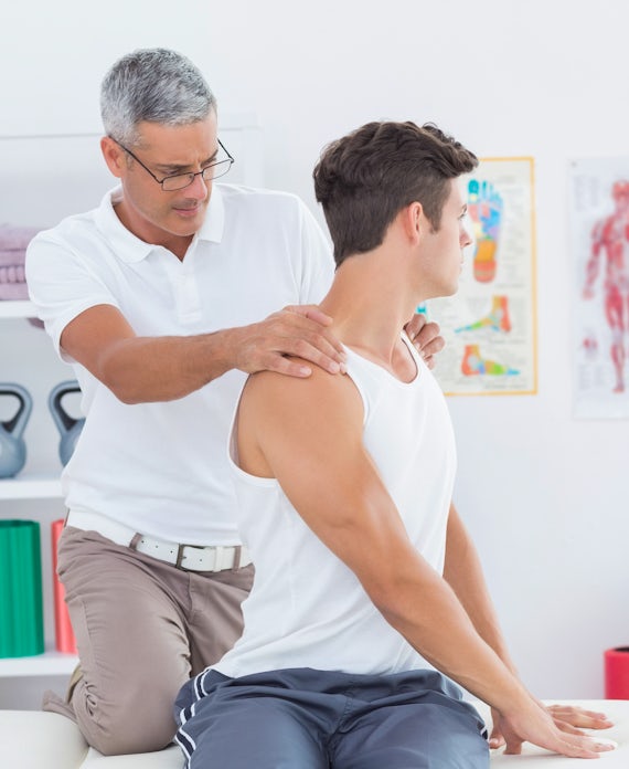 A therapist assists a patient with a shoulder stretch in a bright clinic, surrounded by fitness equipment and anatomical charts.