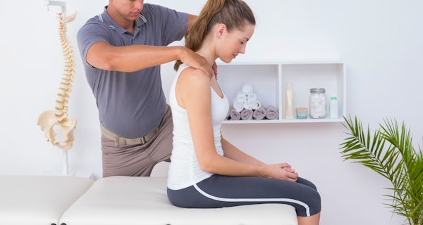 A man is giving a shoulder massage to a woman seated on a treatment table in a bright room with exercise equipment and a spine model.