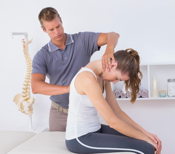 A therapist assisting a woman with neck tension, while she sits on a treatment table in a well-lit, tidy therapy room.