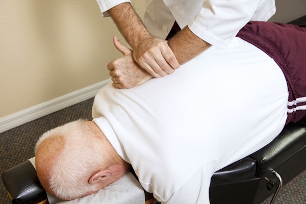 The image shows a chiropractor performing an adjustment on a patient lying face down on a treatment table. The focus is on manual therapy techniques.