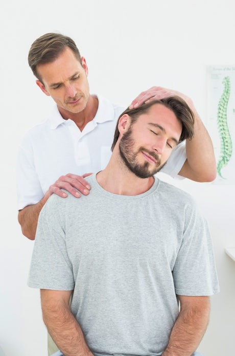 The image shows a man receiving a neck adjustment or massage from a therapist in a bright, clinical setting. Exercise equipment is visible in the background.