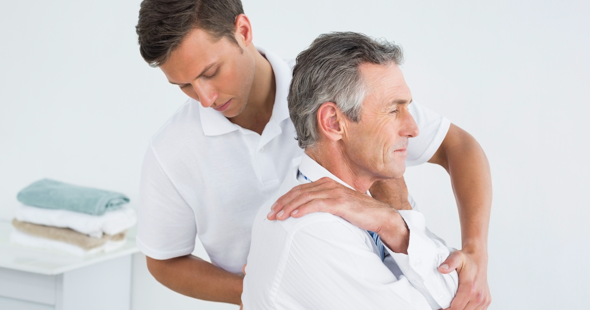 A young man is assisting an older man with a therapeutic shoulder stretch in a bright, clean environment, likely a massage or therapy setting.
