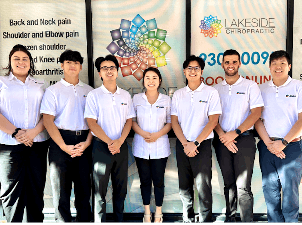 The team at Lakeside Chiropractic standing in front of the clinic window, smiling and wearing white branded uniforms. The background features the Lakeside logo and list of conditions treated, including back, neck, and joint pain.