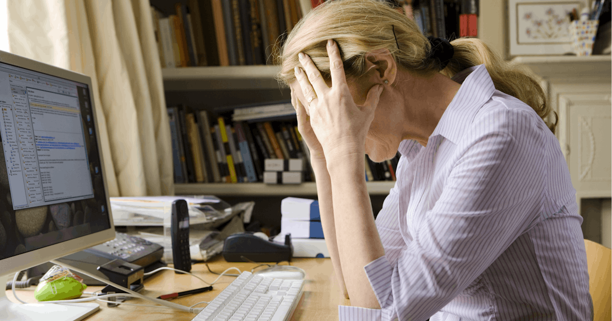 A person sits at a desk with a computer, looking stressed and frustrated, amid papers and a cluttered workspace.