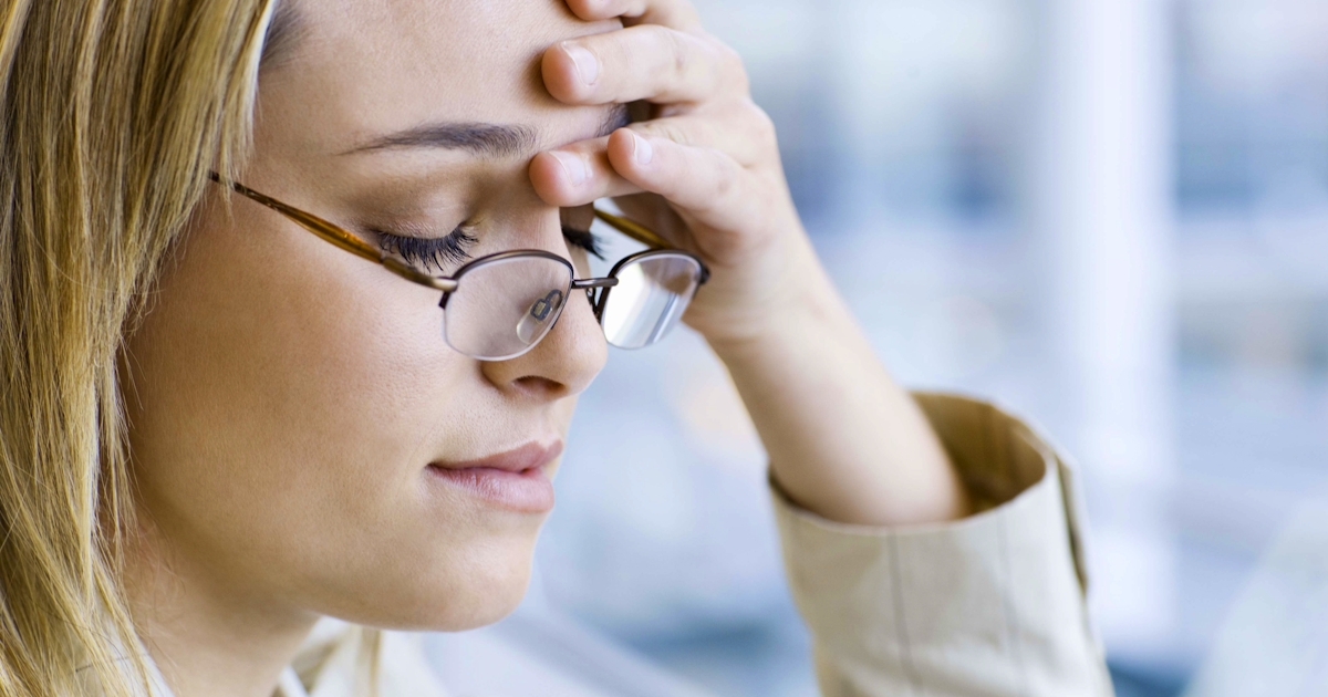 A woman with blonde hair is wearing glasses and resting her forehead on her hand, appearing thoughtful or stressed in a bright indoor setting.