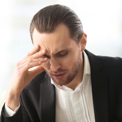 A man in a suit sits at a desk with a laptop, showing signs of stress or concern, while holding his head with one hand. A glass of water is nearby.