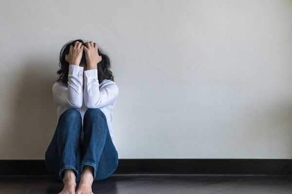 A person sits against a wall, holding their head in their hands, conveying a sense of distress or overwhelm in a minimalistic setting.