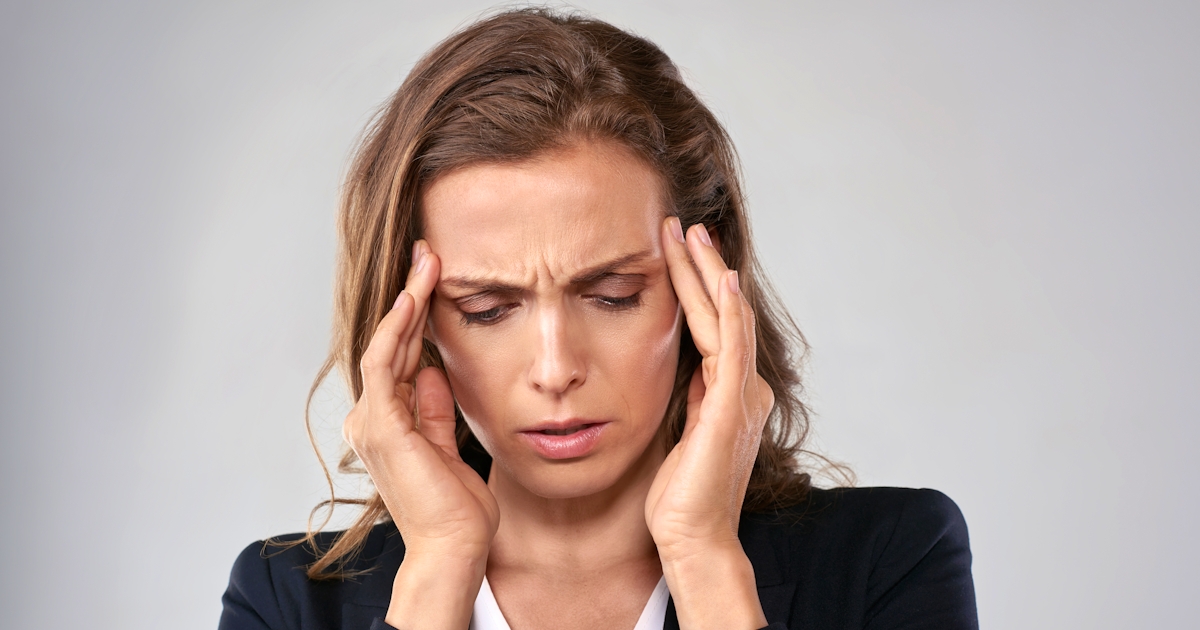 A woman in a suit appears distressed, with her hands on her temples, suggesting she is experiencing stress or a headache.