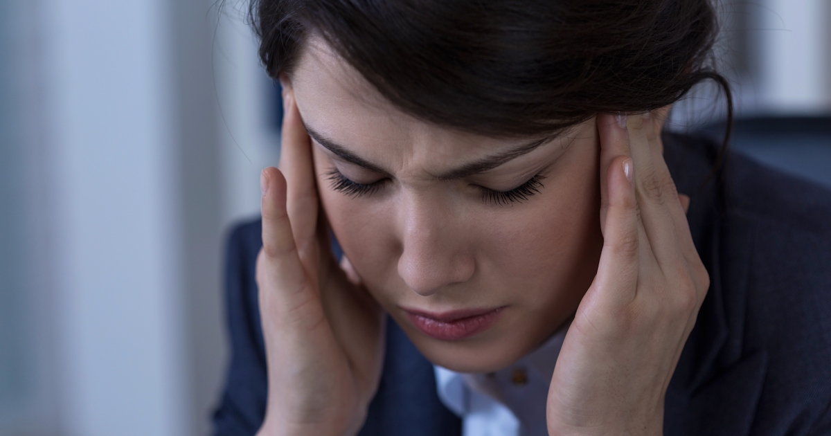 A woman in a suit holds her head with both hands, appearing stressed or in discomfort, possibly indicating a headache or anxiety.