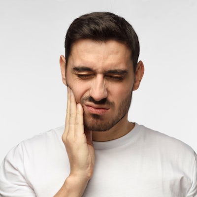 A man is grimacing while holding his jaw, indicating possible pain or discomfort, against a plain background. He is wearing a simple white shirt.