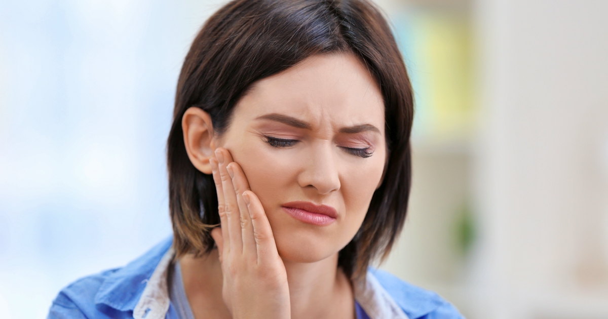 The image shows a woman grimacing in discomfort, holding her jaw, indicating she may be experiencing tooth pain or discomfort.