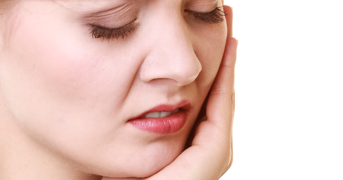A woman holds her jaw with a pained expression, indicating discomfort or distress, against a plain background.