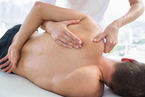 The image shows a person receiving a massage, focusing on the upper back and shoulder area, in a bright, modern setting.