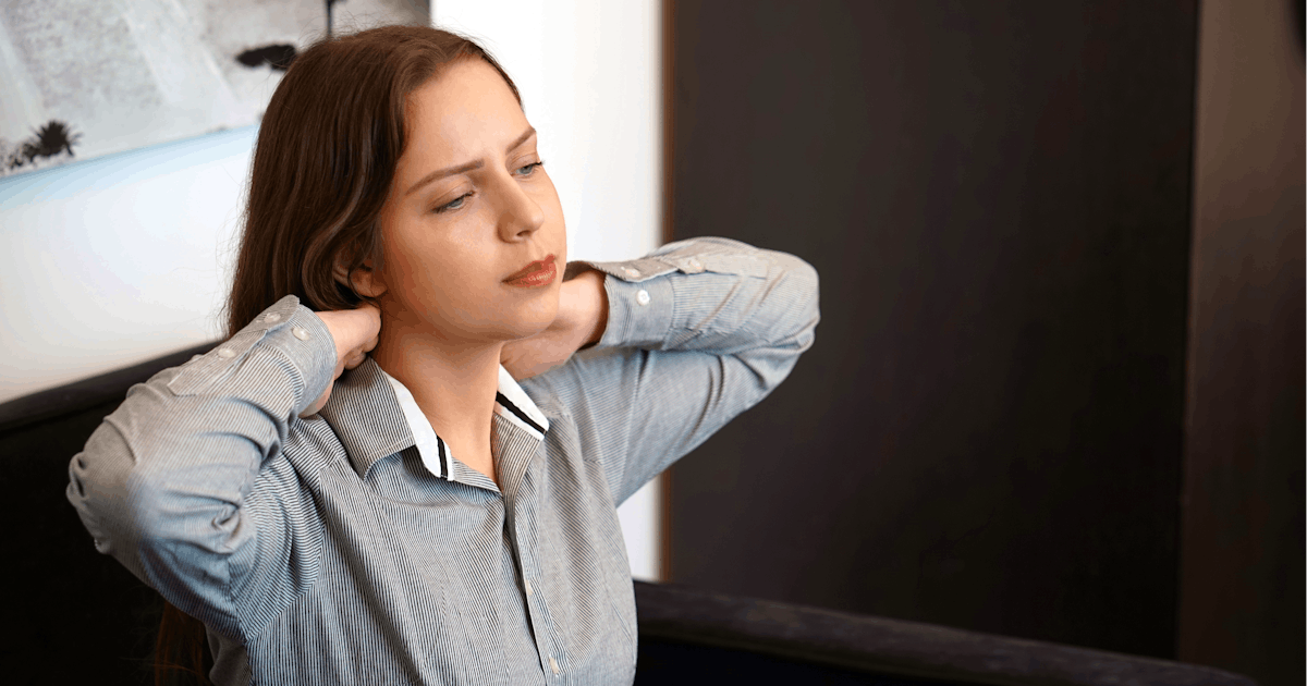 A person with long hair is sitting on a couch, lightly massaging their neck and appearing relaxed or contemplative in a softly lit room.