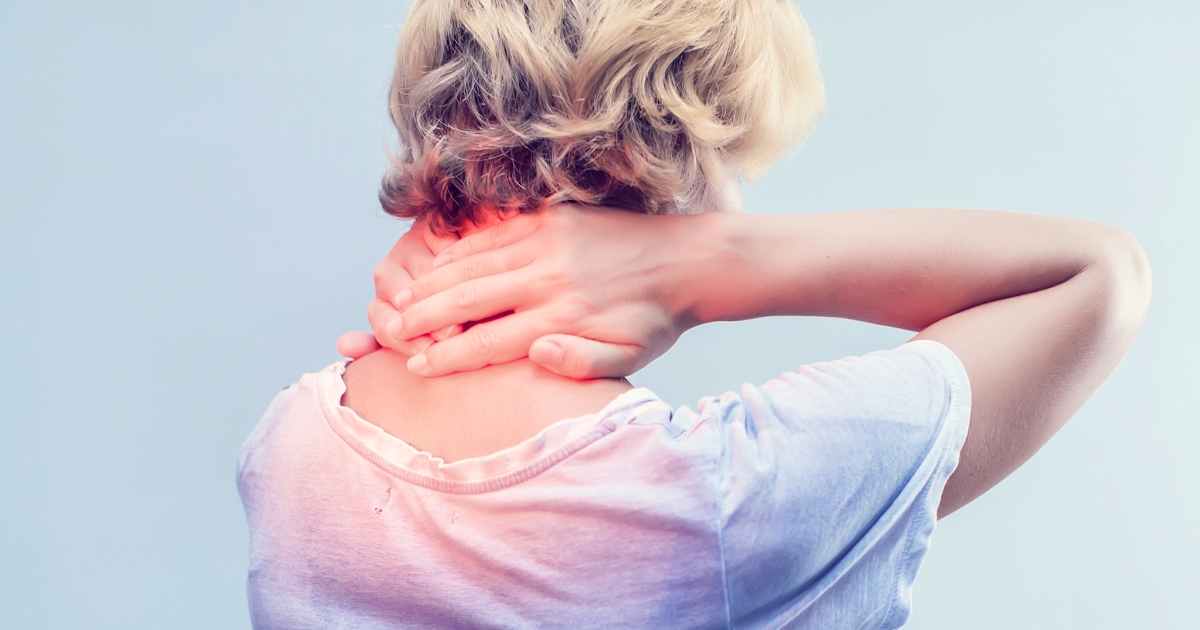 A person with short hair is seen from behind, holding their neck in discomfort while wearing a loose-fitting white shirt against a light background.