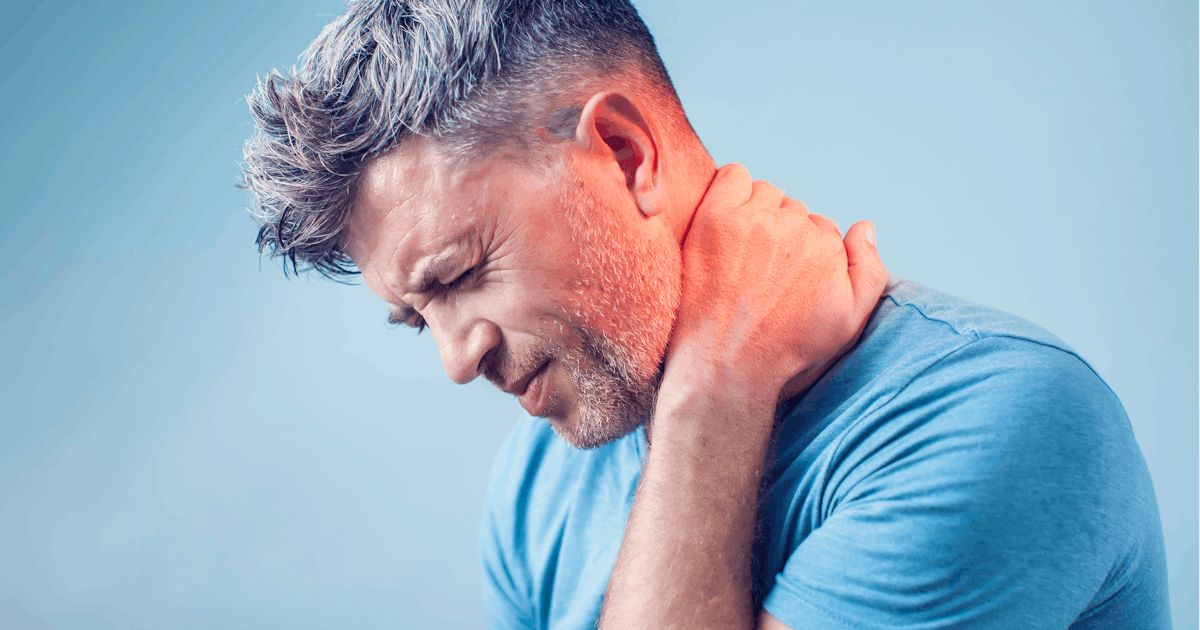 A man is holding his neck in discomfort, showing signs of pain or strain. He has short, styled hair and is wearing a blue shirt against a light background.