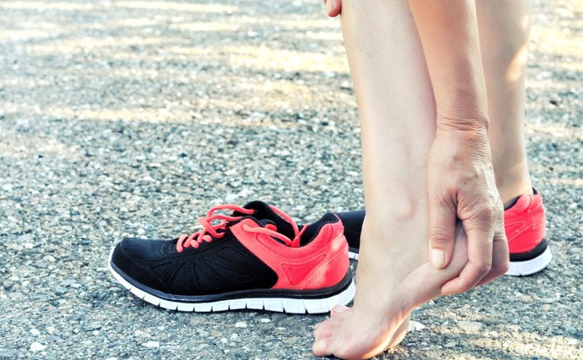 person standing barefoot next to athletic shoes holding their heel in discomfort highlighting how improper or unsupportive footwear can contribute to achilles tendon pain