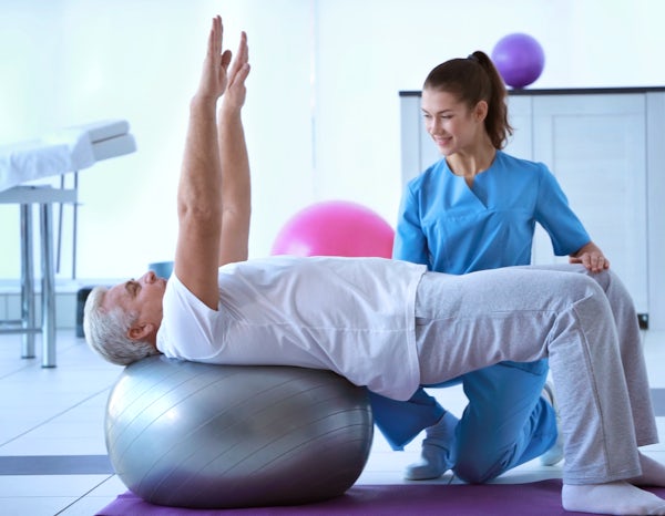 female trainer doctor helping elderly man on exercise ball in studio