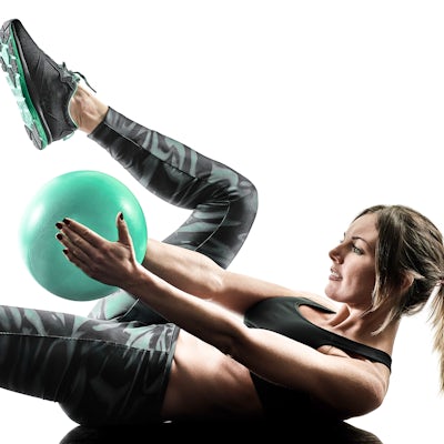 A person is performing an exercise with a light green ball, lying on their back with legs raised, wearing fitness attire and athletic shoes.