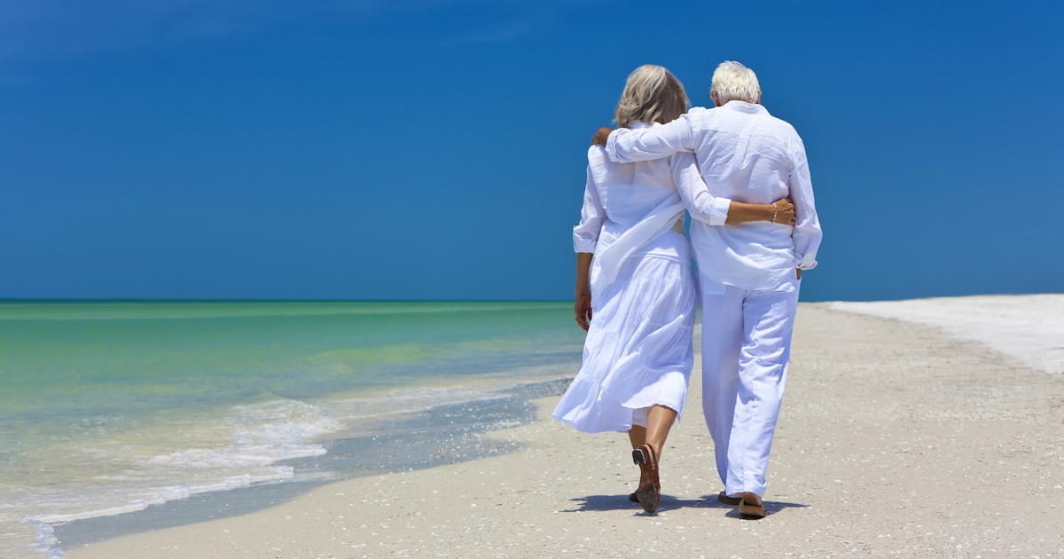 from behind senior couple walking close together on the beach shoreline
