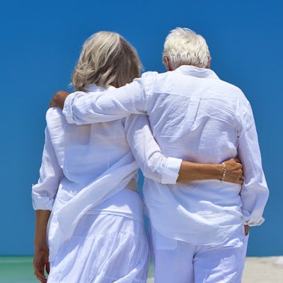 from behind senior couple walking close together on the beach shoreline
