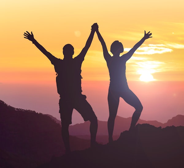 silhouette of two joyful people on mountain top at sunset with arms in the air 