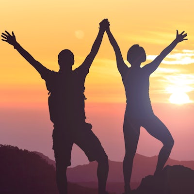 silhouette of two joyful people on mountain top at sunset with arms in the air 