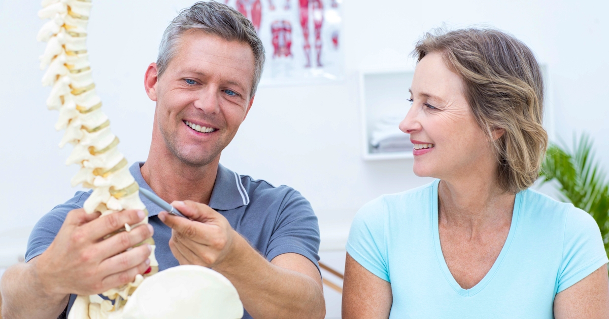 A man is showing a spine model to a woman, who appears engaged and smiling. They are in a clinical or educational setting.