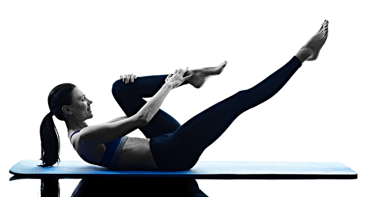 A person performs a pilates exercise on a blue mat, engaging their core with a focus on flexibility and strength. The image is in silhouette.